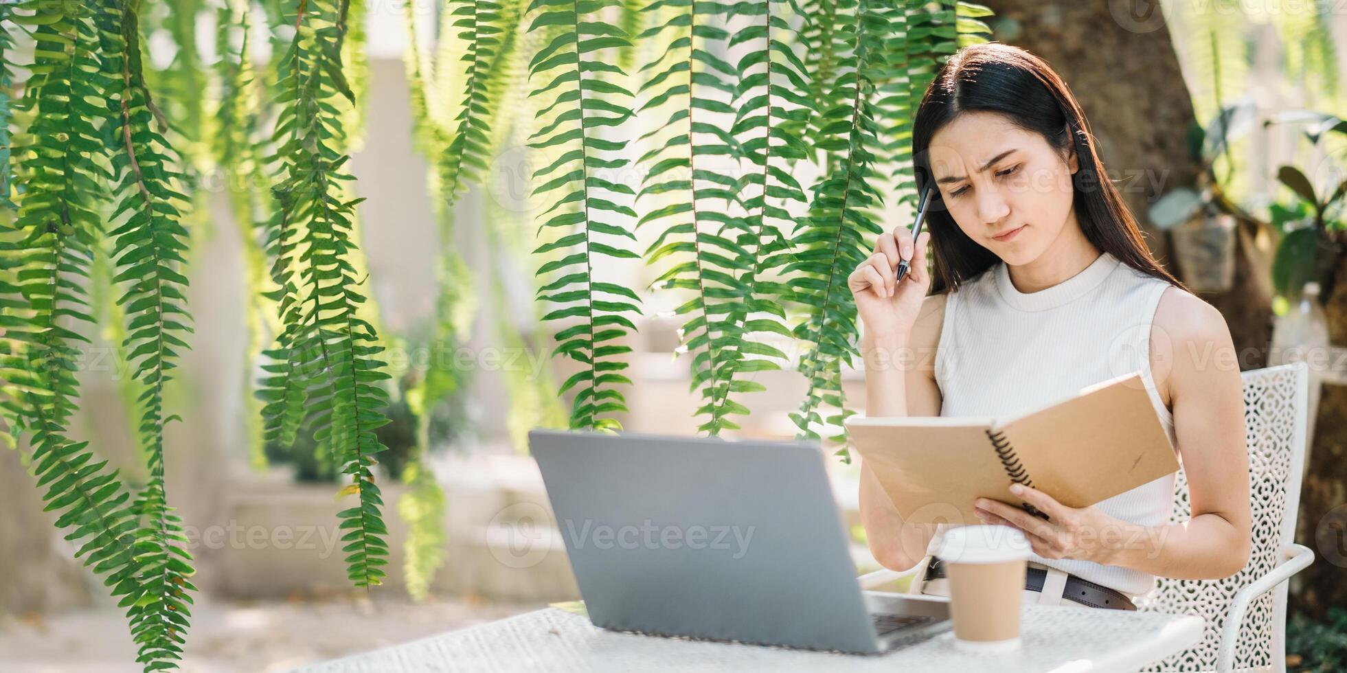 Concentrated young woman reviews her notes, pen to chin, while working on her laptop in a refreshing outdoor setting with foliage. photo