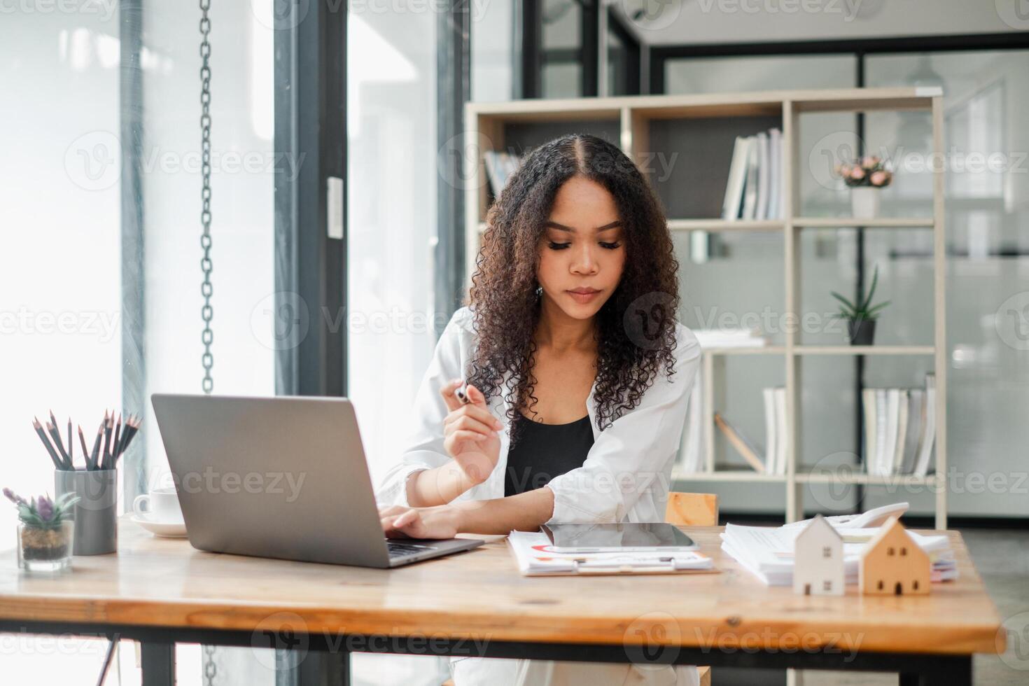 Real estate agent examines property documents while working on a laptop in a modern office, with miniature house models on the table. photo