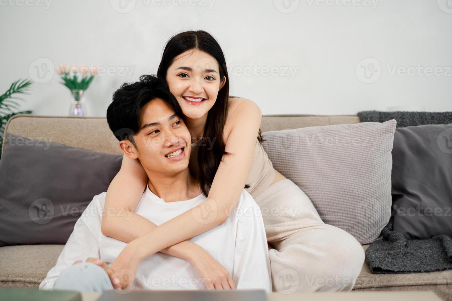 Woman in a sleeveless dress smiles down at a man in a white t-shirt as they embrace on a sofa with a laptop open in front of them. The setting is a cozy living room with decorative pillows and plants. photo