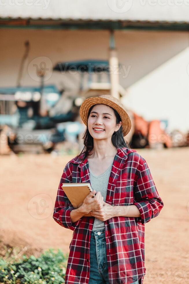 Smiling young farmer holding a notebook stands proudly on her farm with agricultural machinery in the background. photo