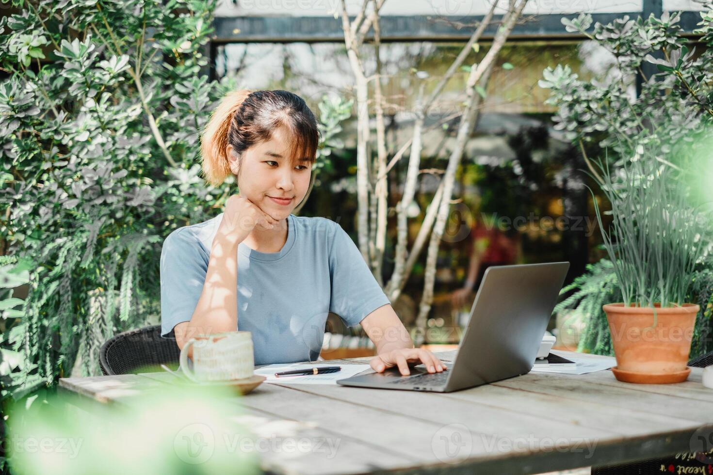 Contemplative woman works on her laptop at a wooden garden table, a serene green setting with plants and sunlight enhancing the calm work environment. photo