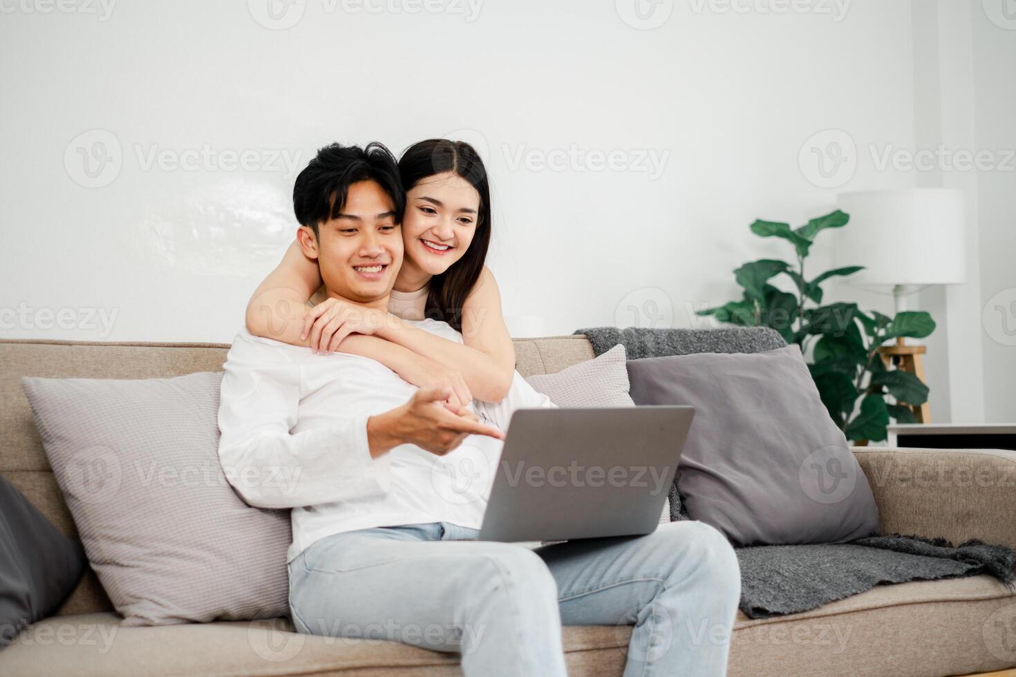 Young Asian couple enjoys a cozy moment on their sofa, interacting with a laptop, in a light-filled, plant-adorned living space. photo