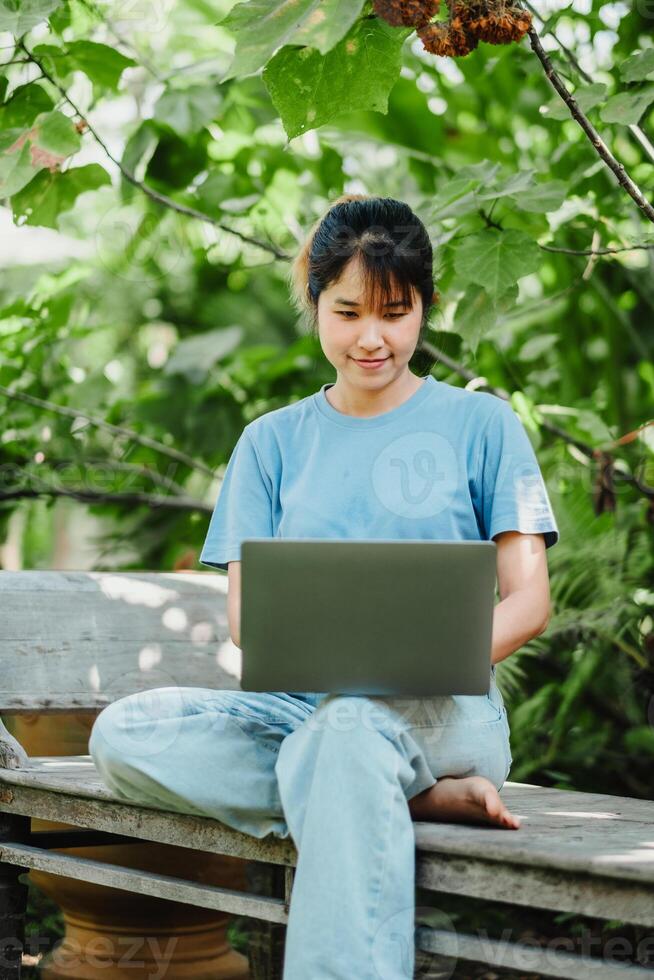 Smiling young woman sitting on a wooden bench, working on her laptop in a lush green garden setting. photo