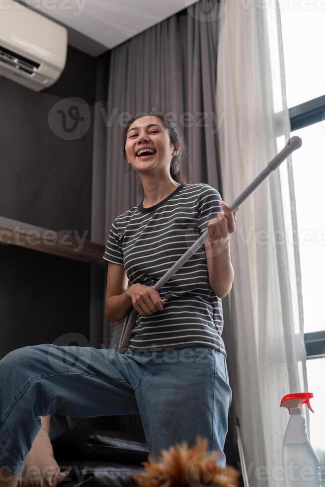 House cleaning with fun. Happy young asian housewife singing song during cleanup, using mop as guitar, enjoying domestic work. Young woman dancing and cleaning in living room photo