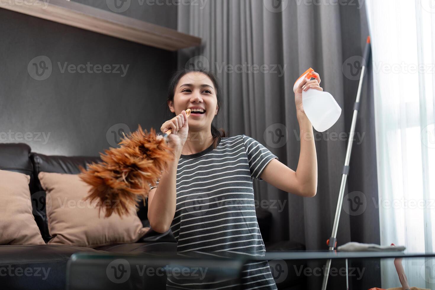 House cleaning with fun. Happy young asian housewife singing song during cleanup, using feather duster as microphone, enjoying domestic work. Young woman dancing and cleaning in living room photo