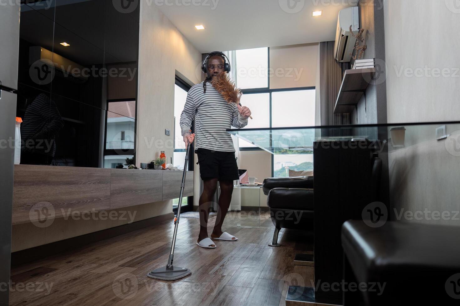 Funny African American is cleaning house doing housework cleaning floor with mop and listening to music in wireless headphones, African American man is dancing and singing photo