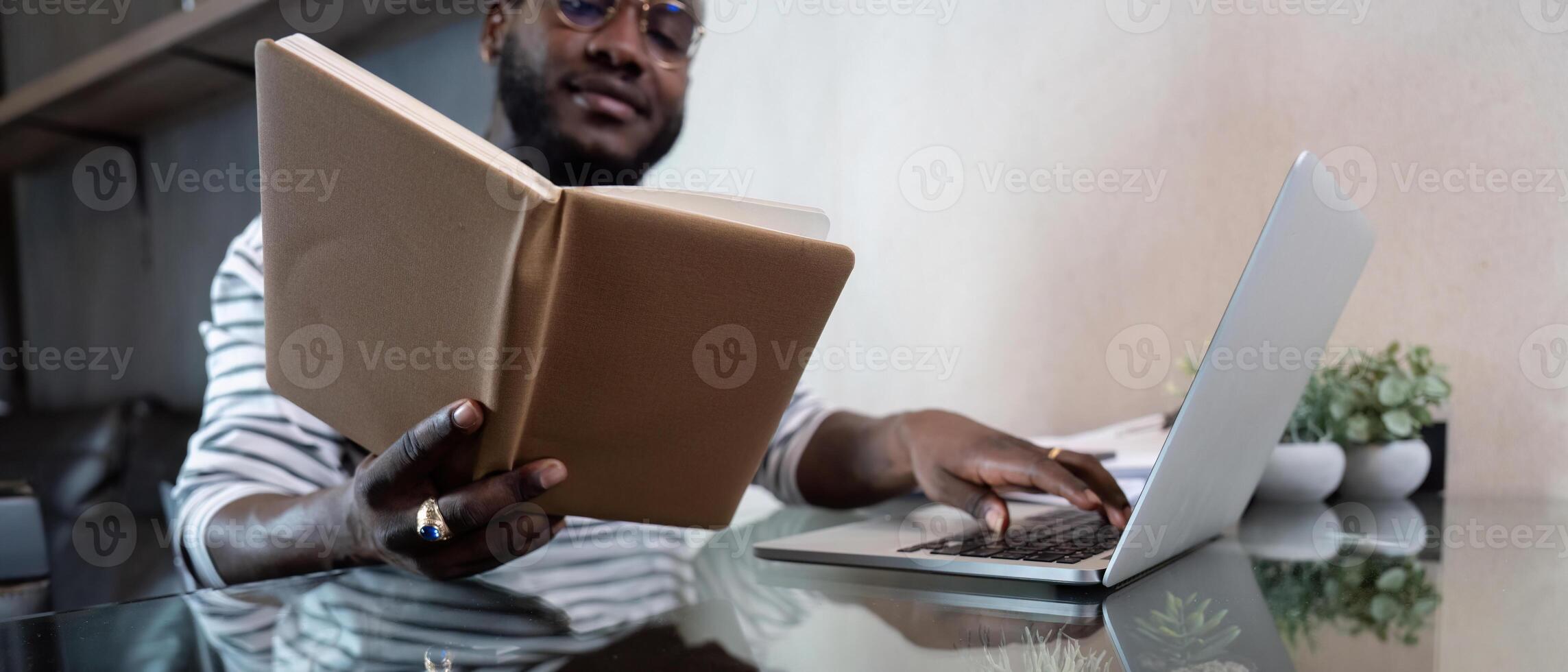 Young african man using laptop at home, black male looking at read book relaxing on leisure with work sit on glass table in living room photo