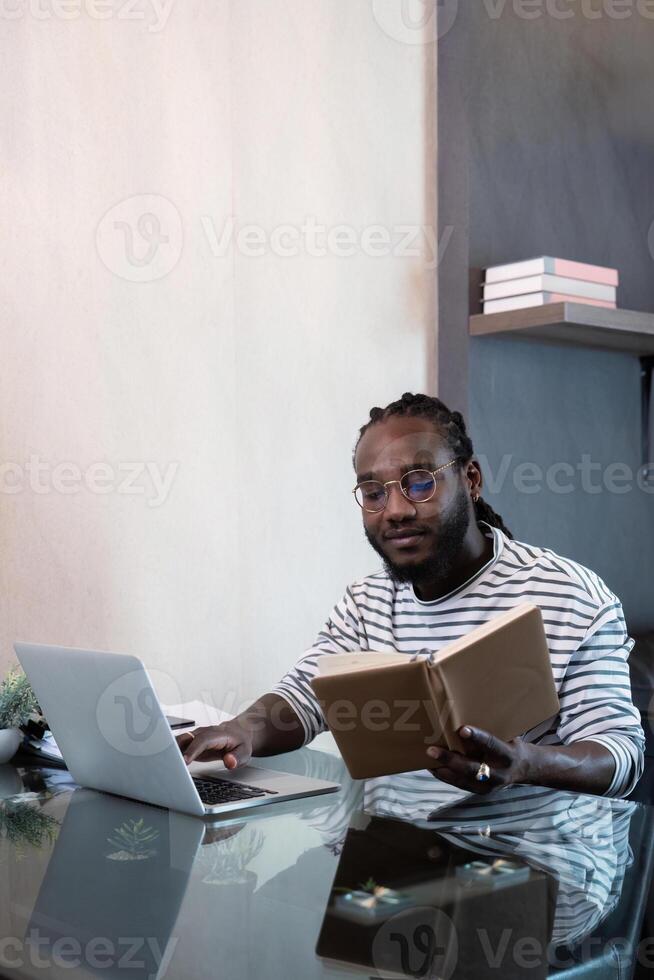 Young african man using laptop at home, black male looking at read book relaxing on leisure with work sit on glass table in living room photo