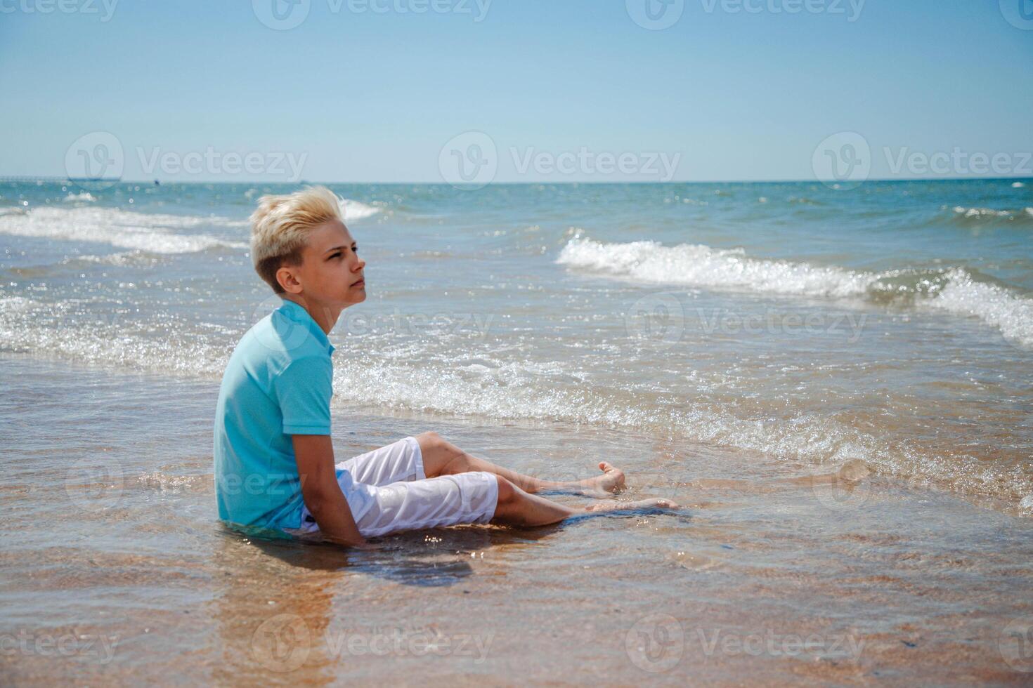 Handsome teenager boy of European appearance with blond hair in white shorts, and a blue T-shirt sits on a beach in sea water and looks thoughtfully into the distance. Summer vocation concept.Summer travel sale concept.Copy space. photo