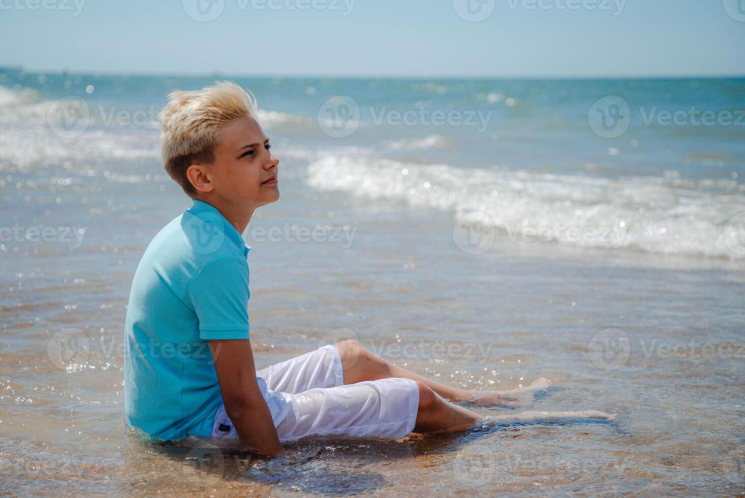 Handsome teenager boy of European appearance with blond hair in white shorts, and a blue T-shirt sits on a beach in sea water and looking away. Summer family vacation concept.Summer travel concept.Copy space. photo