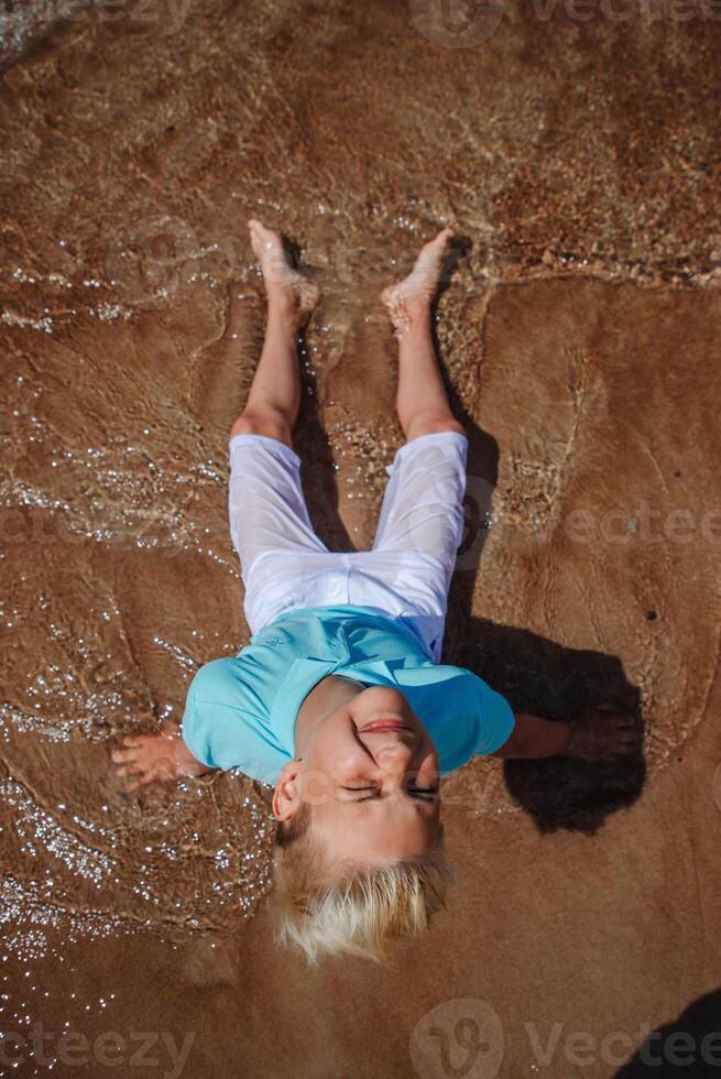 Handsome teenager boy of European appearance with blond hair in white shorts, and a blue T-shirt sits on a beach in sea water and looks to the camera. Summer vocation concept.Summer travel sale concept.Top view photo.Vertical photo. photo