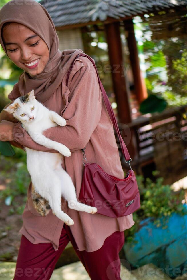 woman wearing a headscarf is holding a white cat on a background of plants as copy space. photo