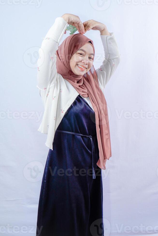 Javanese woman wearing a hijab is showing a sign of love by forming a heart above her head, on a white background with empty space for advertising. photo