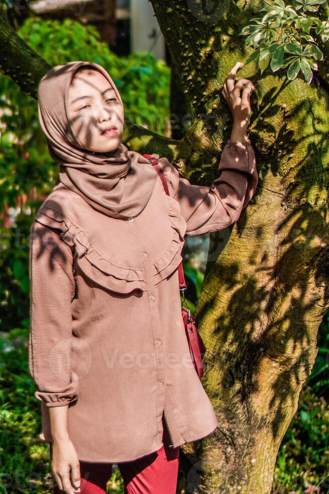 un velado mujer estaba en pie siguiente a un árbol con el sombra de el hojas Derecha en su rostro. foto
