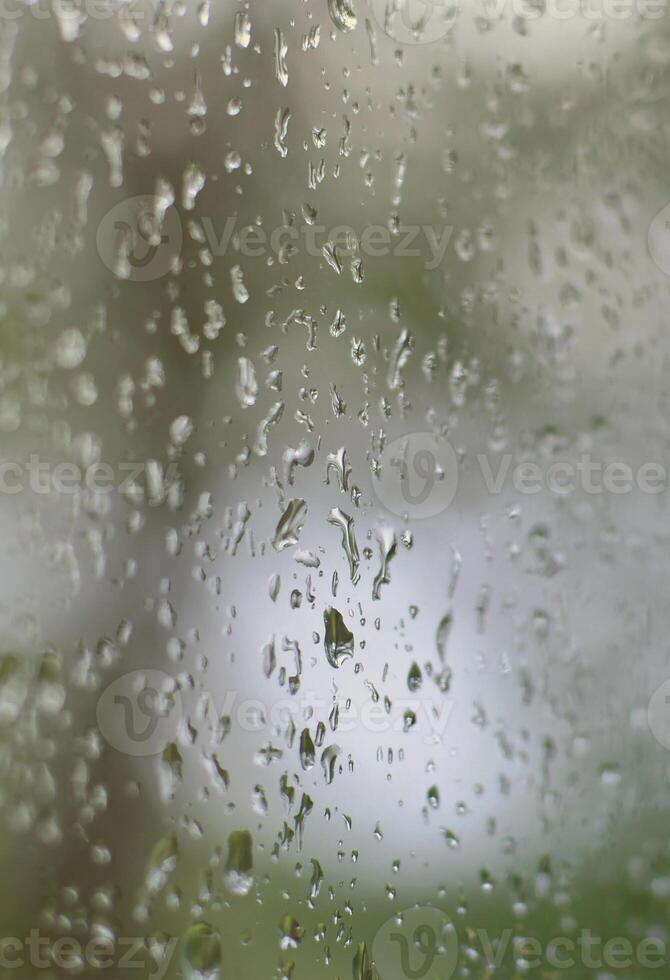 A photo of rain drops on the window glass with a blurred view of the blossoming green trees. Abstract image showing cloudy and rainy weather conditions