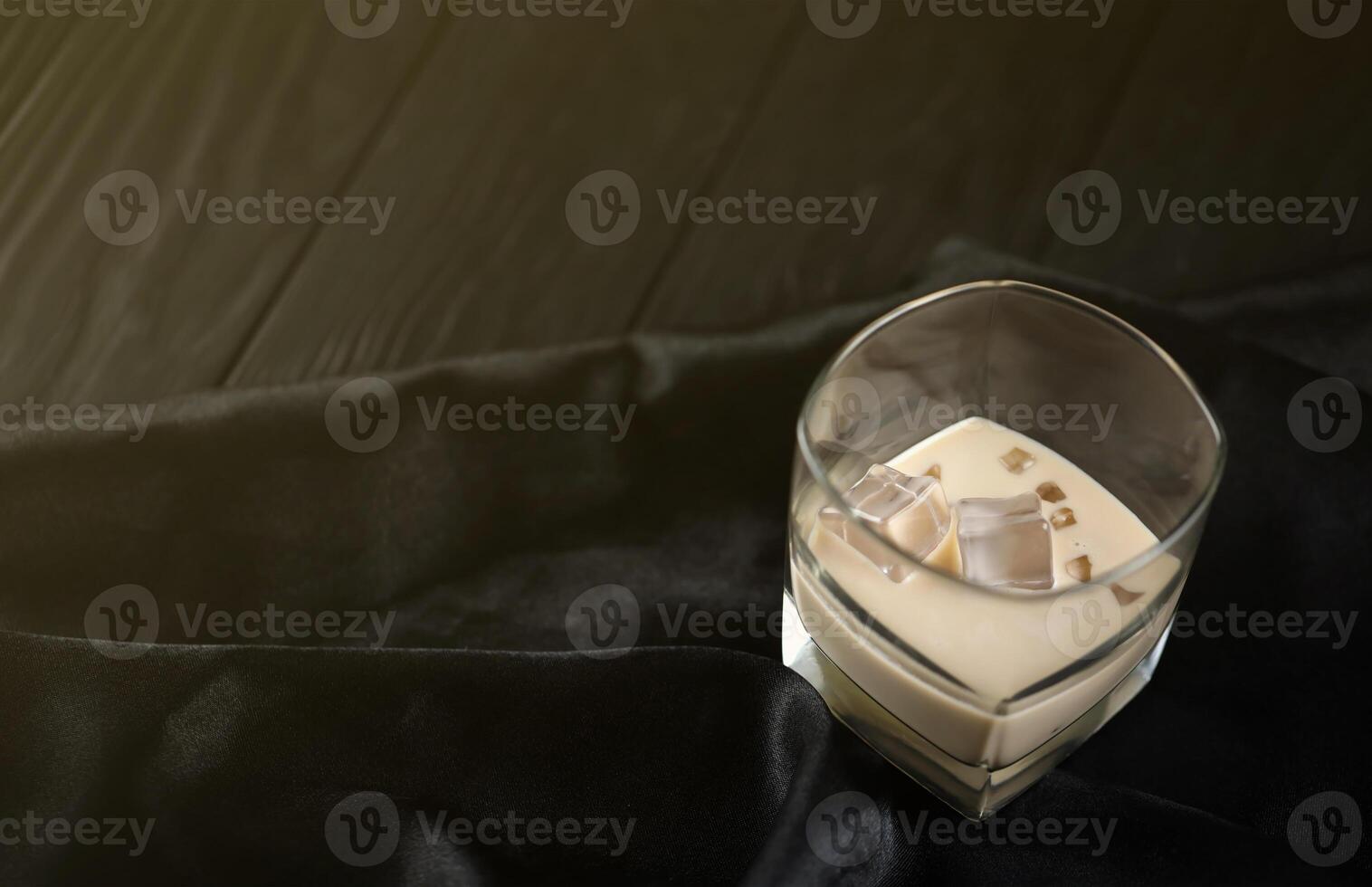 Glass of Irish cream baileys liqueur with ice cubes on dark fabric and wooden bar table background. Homemade luxury elite alcohol cocktail. Place for text photo
