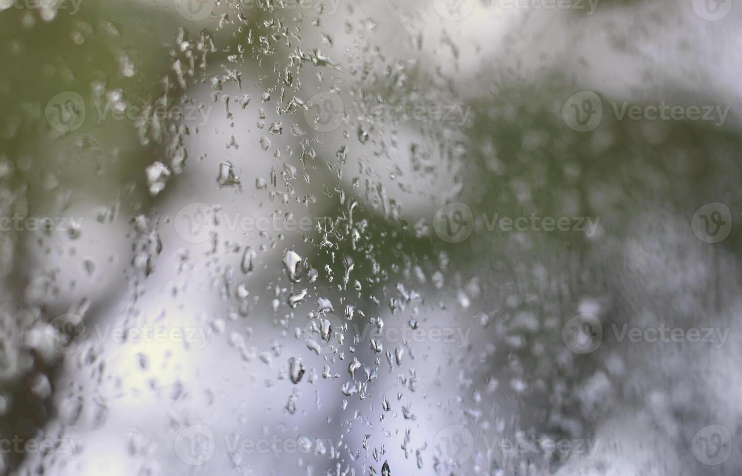 A photo of rain drops on the window glass with a blurred view of the blossoming green trees. Abstract image showing cloudy and rainy weather conditions