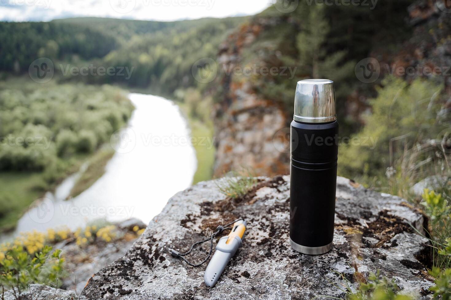 A thermos stands on a stone against the background of the river, a landscape in nature in the mountains, a knife lies on a stone, the concept of a summer holiday in the mountains, hiking, hiking. photo