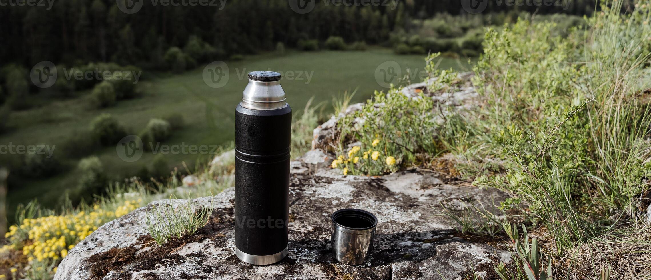 A thermos of tea stands on a mountain against the background of a forest, a landscape of a summer day, a place for a logo, a vacation in the mountains, a mountaineering camp high on top. photo