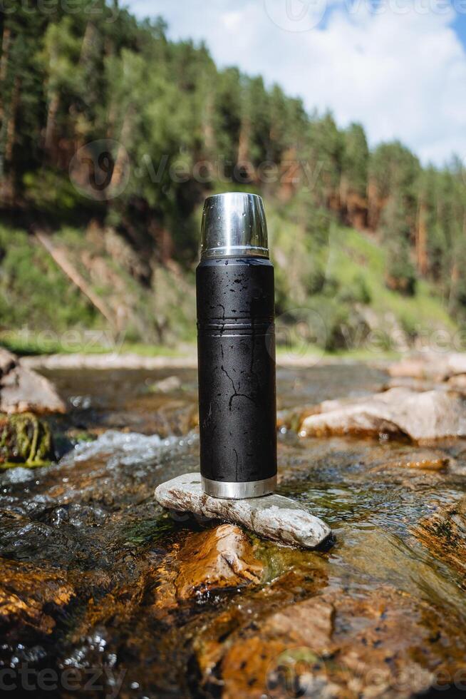 A black metal thermos stands on a stone in a mountain river, a vacuum bottle for hot drinks, tea on a hike in the mountains, a thermo flask. photo