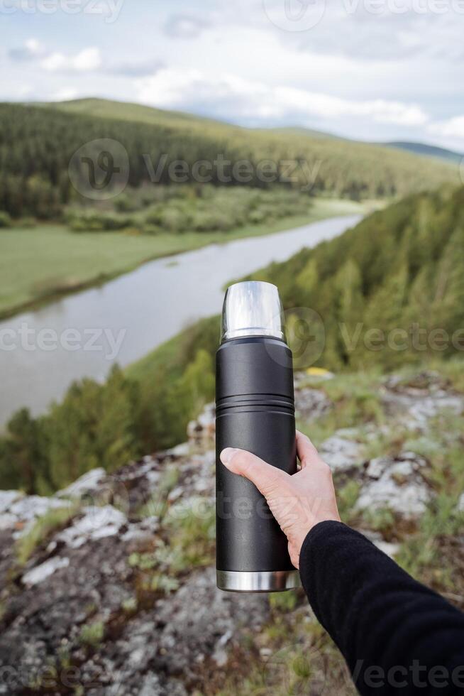 A person's hand holds a metal thermos with tea against the background of nature, a tourist shows a bottle with a hot drink, a black steel thermos. photo