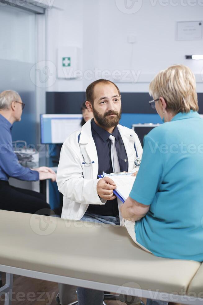 Elderly woman fills out medical forms and talks with doctor about her treatment. Portrait of male practitioner asking senior patient to sign documents for physiotherapy treatment. photo