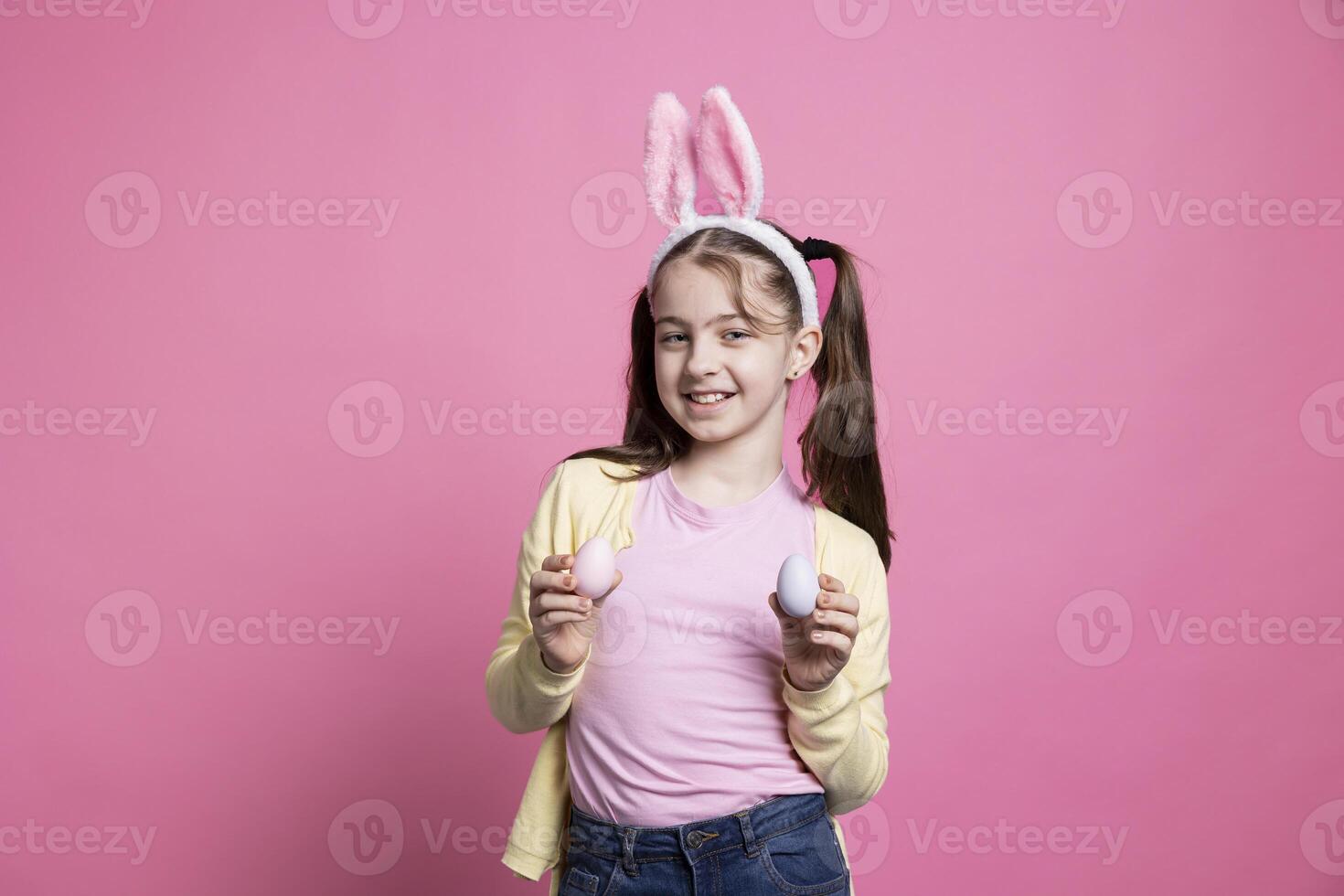 Young toddler smiling and wearing bunny ears in front of camera, presenting her handmade painted pink eggs for easter holiday tradition. Cheerful cute girl in studio shows ornaments. photo