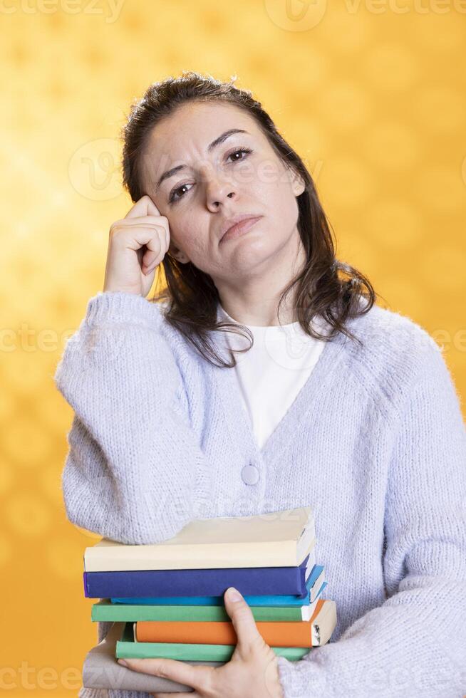 Portrait of pensive woman carrying stack of books, memorizing information for school exam, studio background. Student holding textbooks, remembering and practicing before university assessment photo