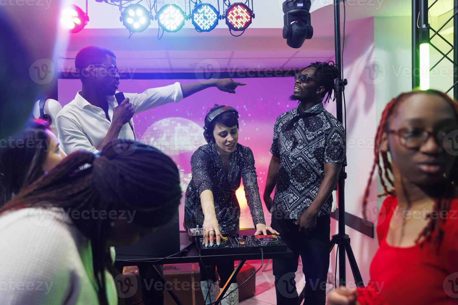 Musicians blending tracks with music mixer console and singing on stage in nightclub. African american men and caucasian woman band playing at disco party in crowded club photo