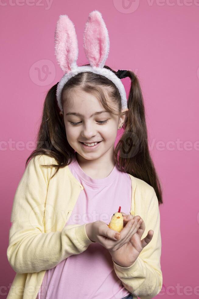 pequeño joven niño demostración un linda relleno polluelo juguete en estudio, alegre pequeño niña con dorado arreglo terminado rosado antecedentes. joven con dulce mullido orejas para Pascua de Resurrección festividad en frente de cámara. foto