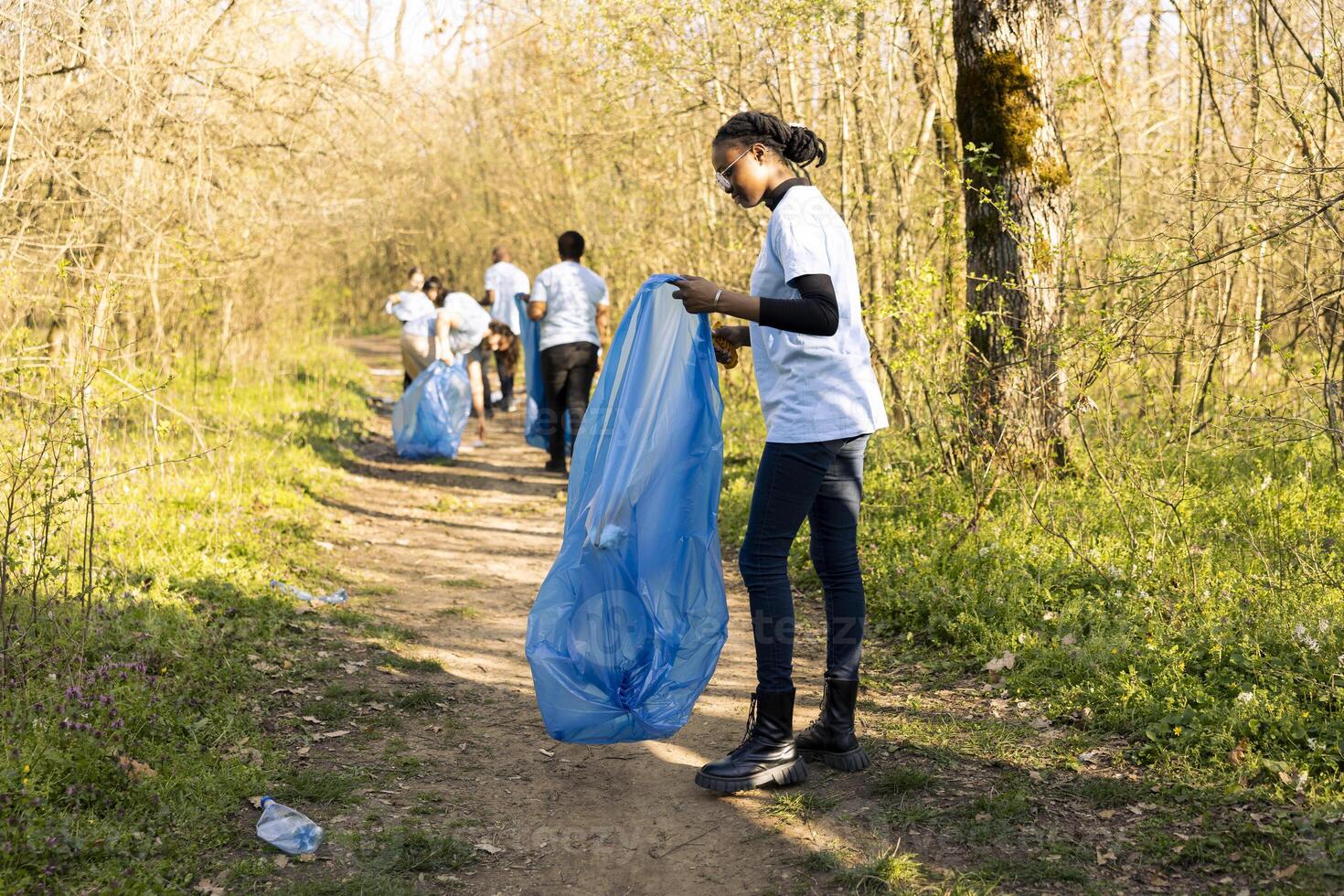 African american girl collecting garbage in a disposal blue bag, supporting nature preservation and helping protect the environment. Proud teenager volunteering to pick up trash. photo