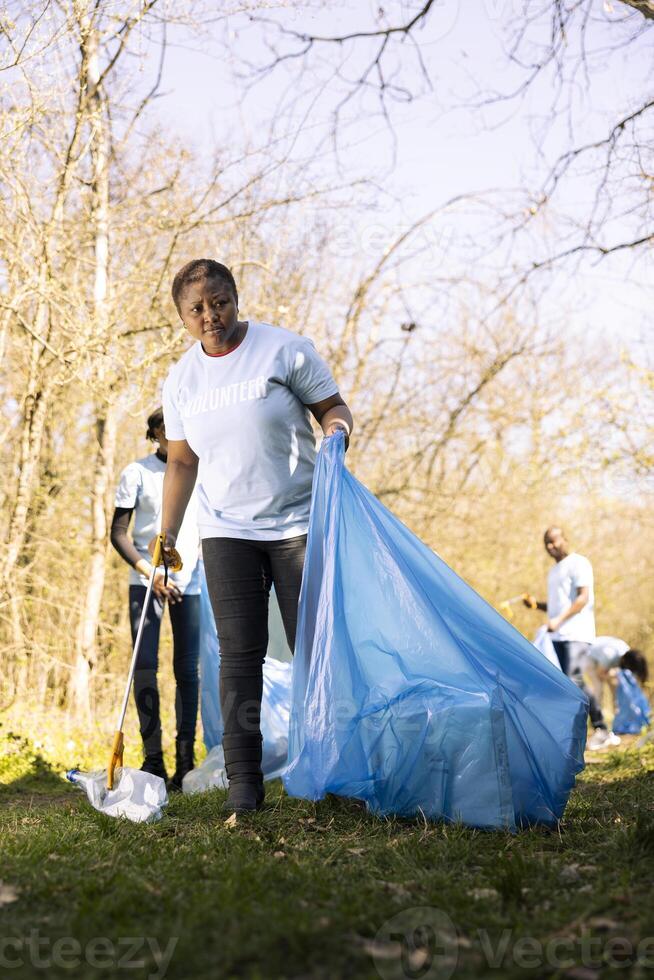 African american volunteer tidying the woods area of garbage and plastic bottles, collecting trash with claw and bags. Young woman doing voluntary work to conserve natural habitat. photo