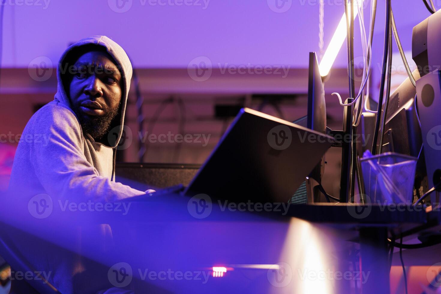 Concerned hacker looking over shoulder, afraid of cyber police while doing cyberattack. Scared african american cybercriminal in hood coding illegal ransomware on laptop in abandonded warehouse photo