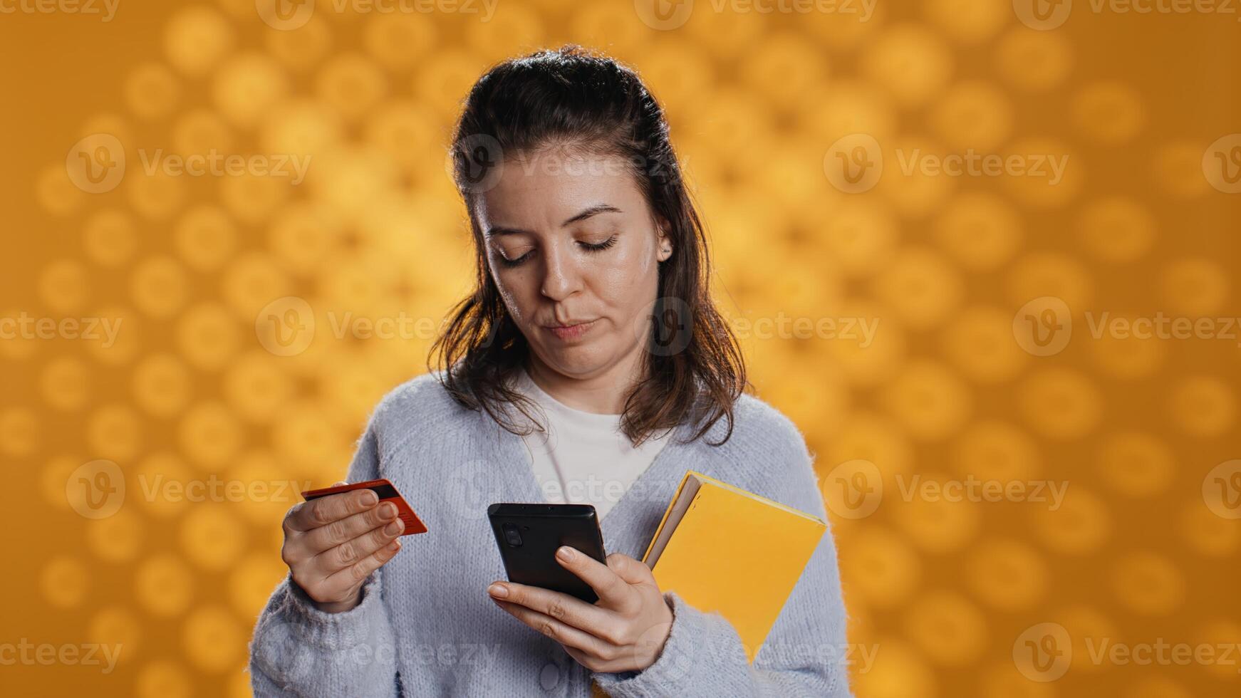 Woman holding books, purchasing more with phone, conveying joy of reading concept, studio background. Person buying novels online with credit card, showing appreciation for literature, camera B photo