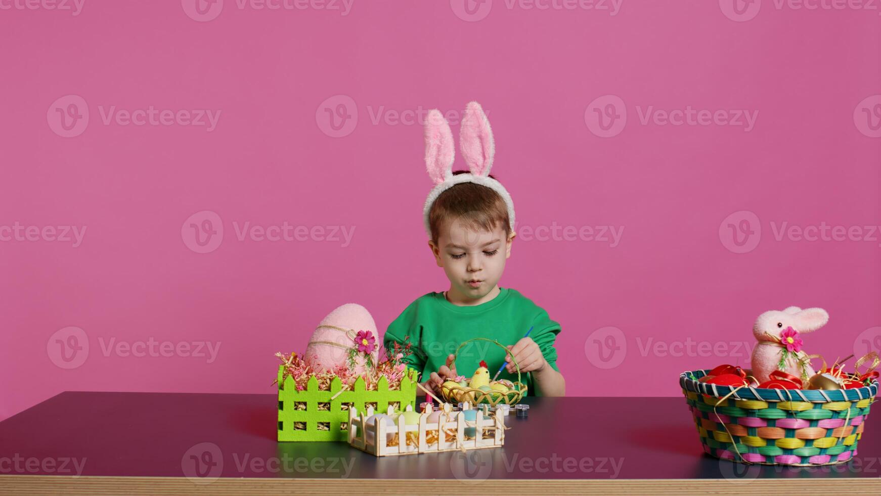 contento pequeño niño arreglando huevos y polluelo en un cesta en preparación para Pascua de Resurrección domingo celebraciones, creando festivo decoraciones en estudio. pequeño chico teniendo divertido colorante. cámara b. foto