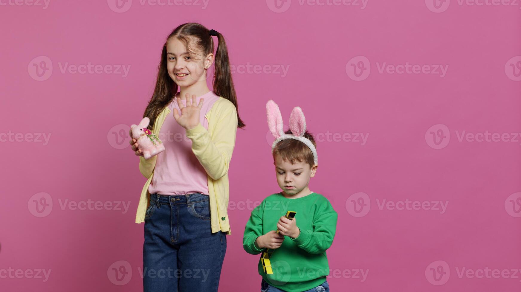 Cute brother and sister posing against pink background in studio, wearing bunny ears and playing with toys. Cheerful siblings feeling excited about easter, traditional spring holiday. Camera A. photo