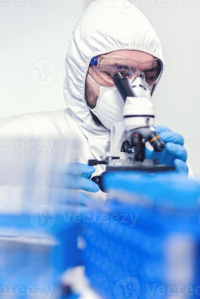 hombre laborante en ppe traje utilizando microscopio haciendo investigación de coronavirus científico en protector traje sentado a lugar de trabajo utilizando moderno médico tecnología durante global epidemia. foto