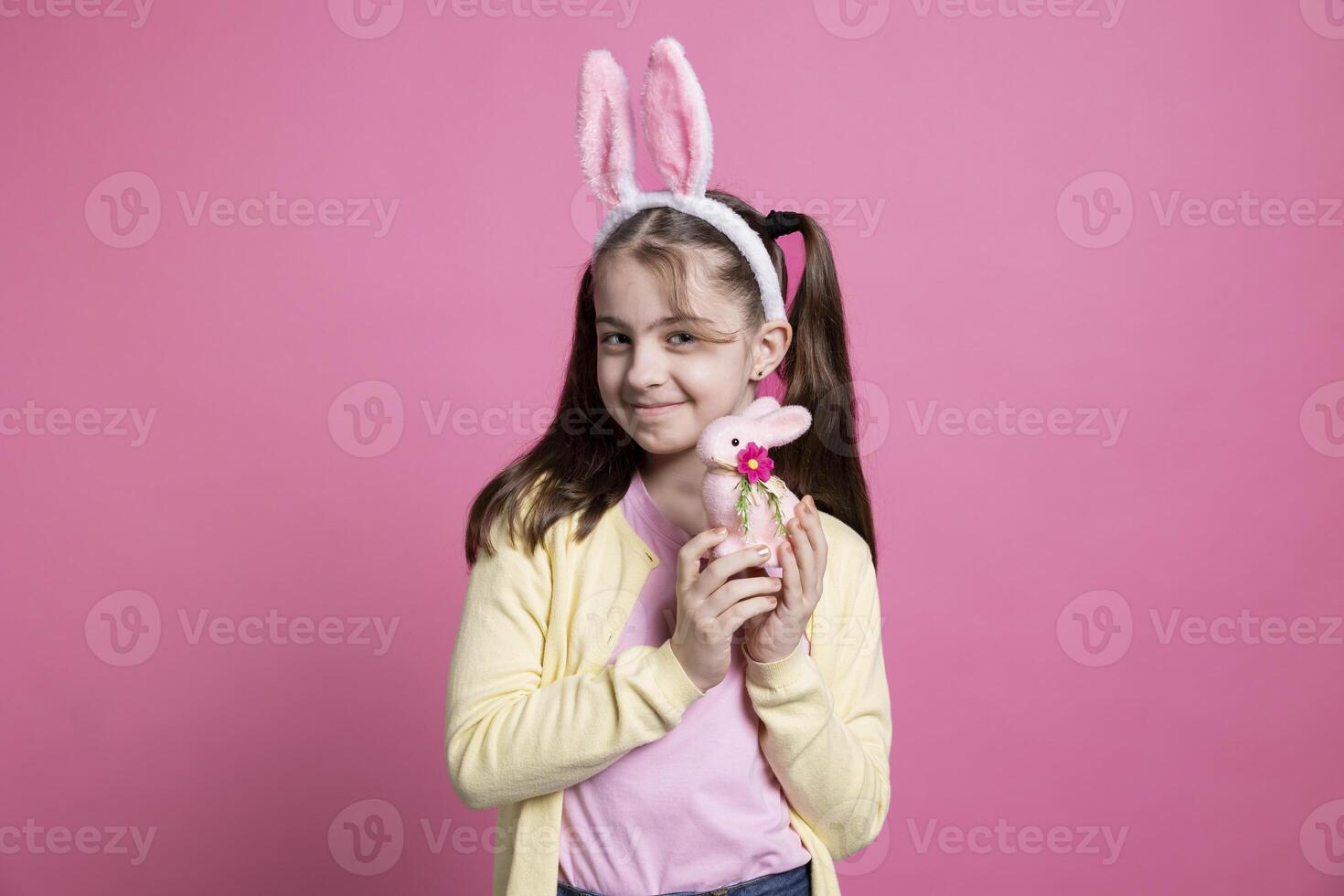 Positive cheerful girl with bunny ears posing over pink background, feeling enthusiastic about easter festive celebration. Cute small child holding a pink rabbit toy in front of camera, fluffy items. photo