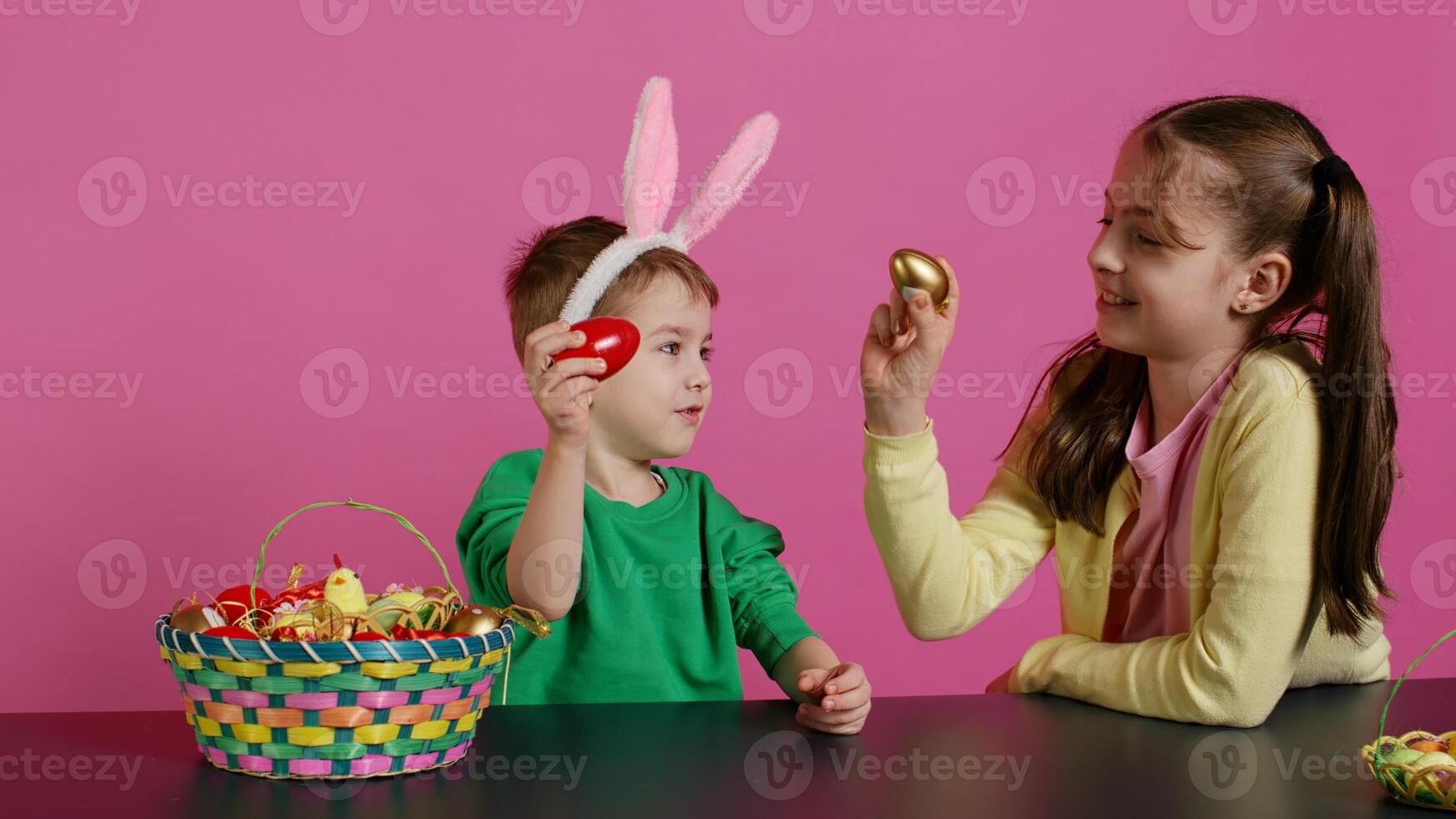 Sweet children knocking eggs together for easter tradition in studio, playing a seasonal holiday game against pink background. Lovely adorable kids having fun with festive decorations. Camera B. photo