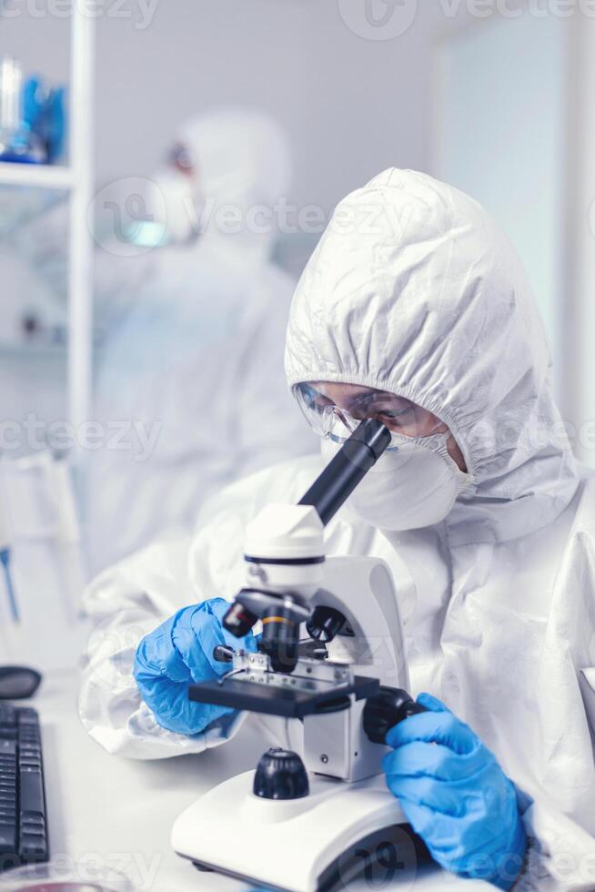 Close up of coronavirus scientist checking vaccine evolution dressed in ppe using microscope. Chemist researcher during global pandemic with covid-19 checking sample in biochemistry lab photo