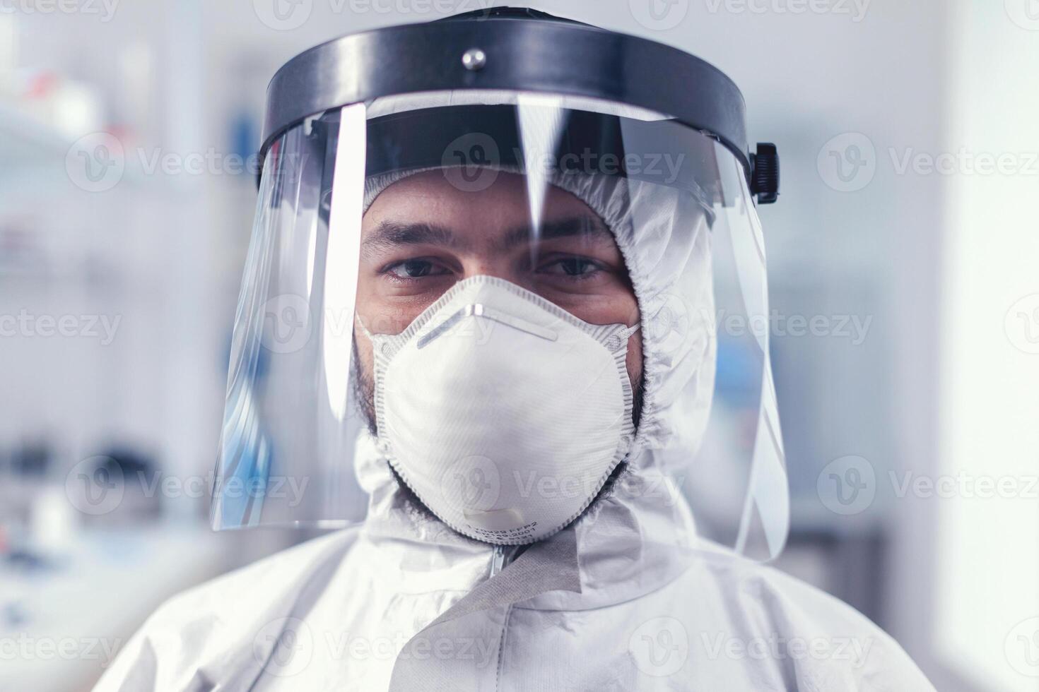 Tired healthcare scientist loooking at camera wearing ppe suit with face shieldin lab. Overworked researcher dressed in protective suit against invection with coronavirus during global epidemic. photo