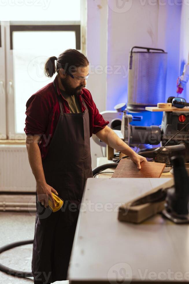 Artisan uses safety goggles to prevent workplace accidents while removing imperfections on wood. Man wearing protective equipment to avoid injury while using sandpaper to do sander plank photo