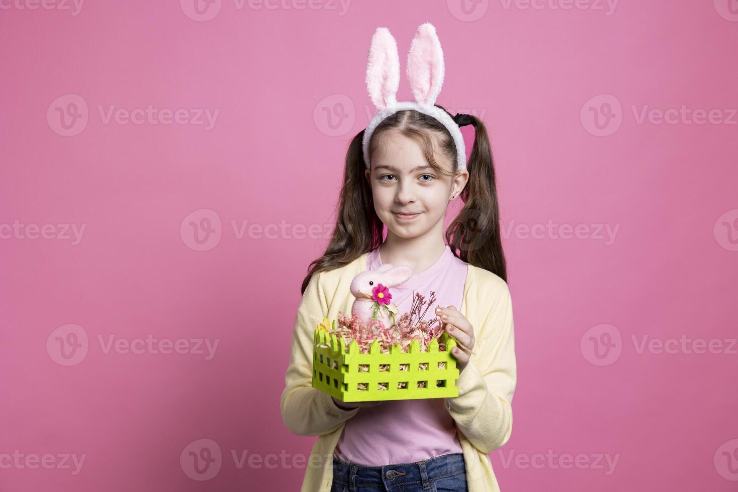 Young optimistic girl showing handmade basket decorated with eggs and a rabbit for easter festivity, posing over pink background. Little cute child with bunny ears presenting spring ornaments. photo