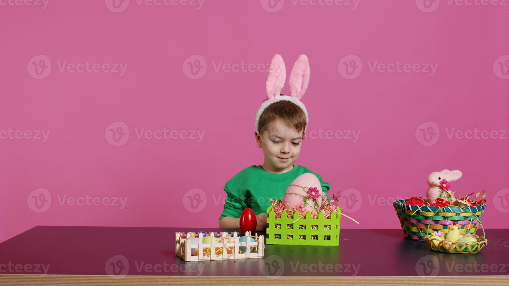 Joyful smiling boy decorating baskets and arrangements for easter sunday celebration, putting plastic grass and fake flowers to craft festive decorations. Happy toddler with bunny ears. Camera B. photo