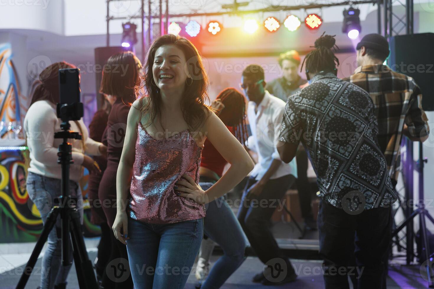 Cheerful smiling woman taking photo on smartphone while having fun in nightclub. Carefree laughing girl recording using mobile phone on tripod while partying in club