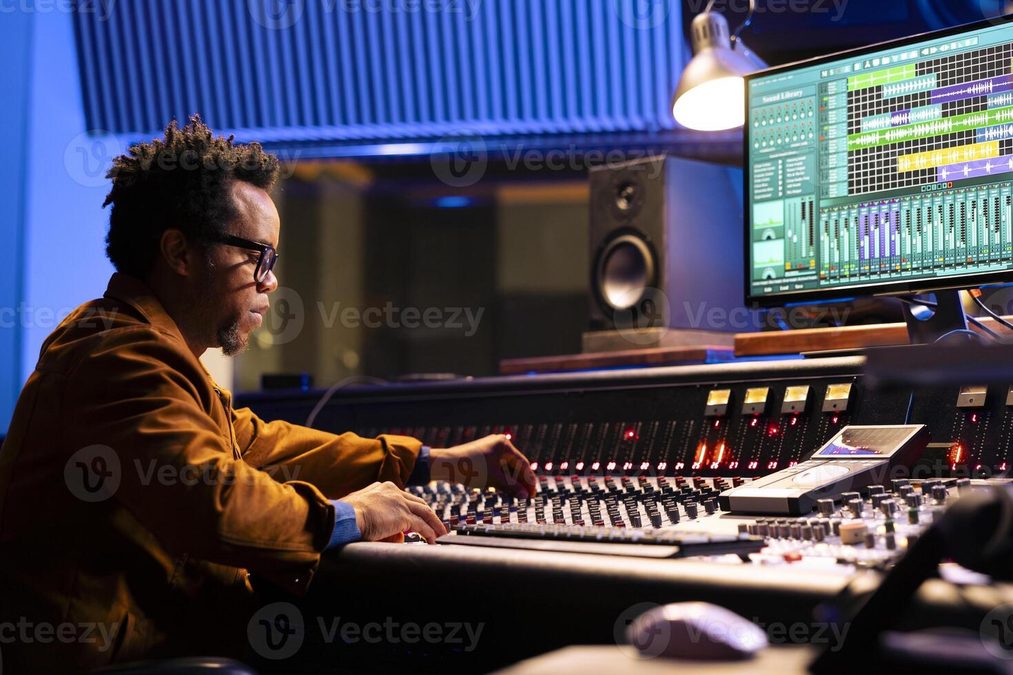 African american sound engineer works on mixing console with sliders and knobs, looking at digital software to record and process tunes. Audio technician creating new music, control room board. photo