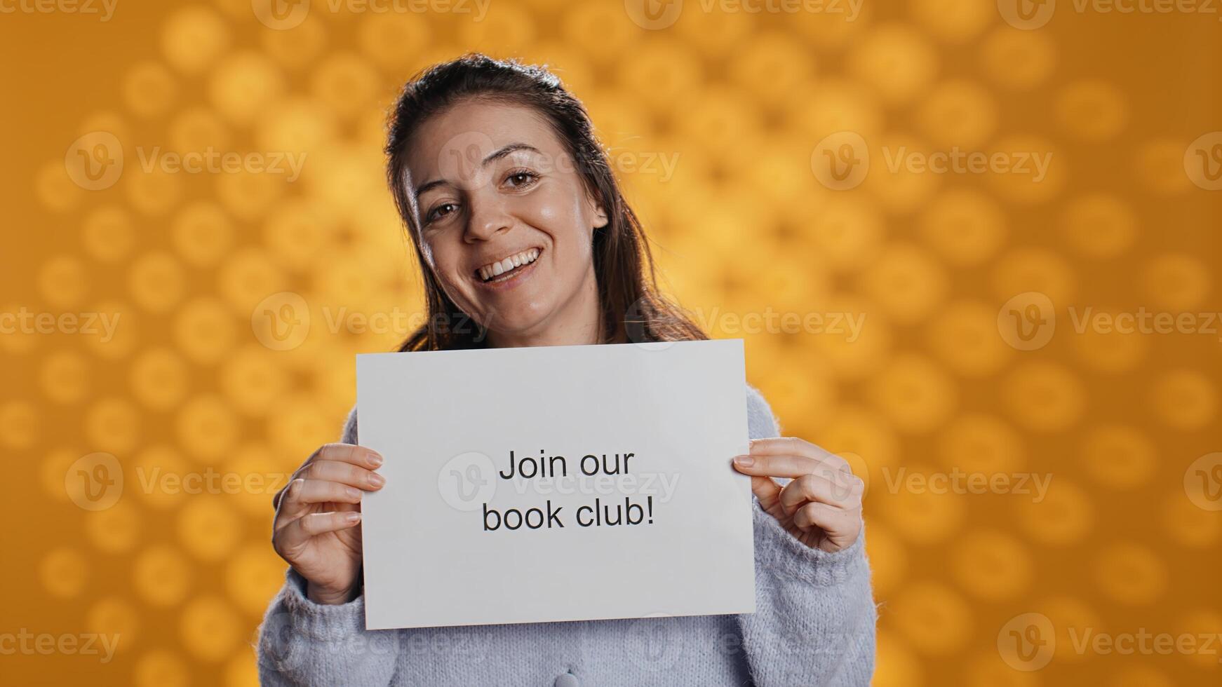 Cheerful book club president holds message urging people to join them, talking about importance of lecture, studio background. Smiling woman inviting bookworms to enlist in her association, camera B photo