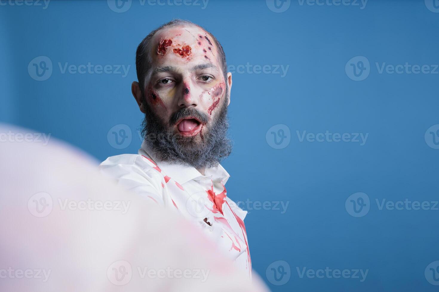 Portrait of brainless zombie isolated over blue studio background doing selfies with tongue out. Mindless undead creature covered in blood and wounds taking pictures with selfie camera photo