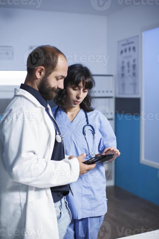 Male doctor and female nurse, together looking at patient files on digital tablet in the medical clinic office. Professional physicians using modern device that holds healthcare information. photo