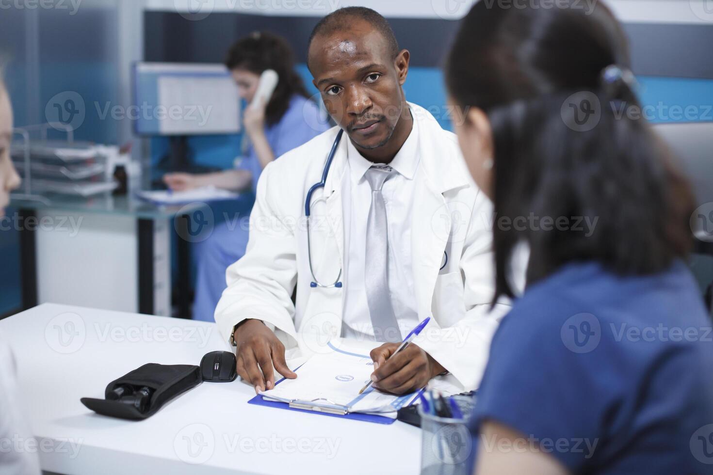 Black man wearing a lab coat, taking notes during a consultation with a female patient at the clinic. African American doctor explains the treatment plan, ensuring proper care. photo